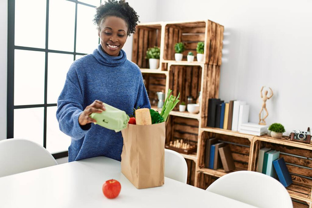 African american woman smiling confident holding food of groceries bag at home - Foto, Bild