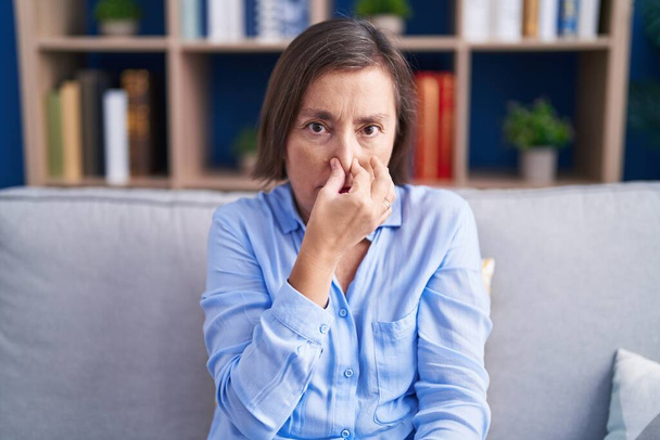 Middle age hispanic woman sitting on the sofa at home smelling something stinky and disgusting, intolerable smell, holding breath with fingers on nose. bad smell  - 写真・画像