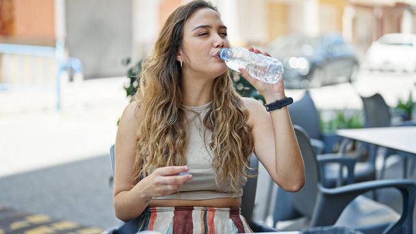 Молодая красивая латиноамериканка пьет воду, сидя на столе на террасе кафе - Фото, изображение