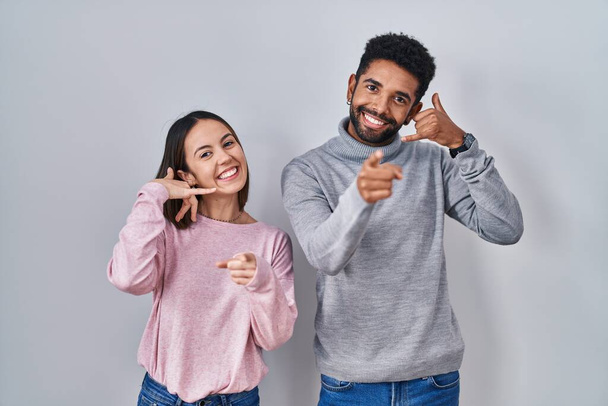 Young hispanic couple standing together smiling doing talking on the telephone gesture and pointing to you. call me.  - Photo, Image