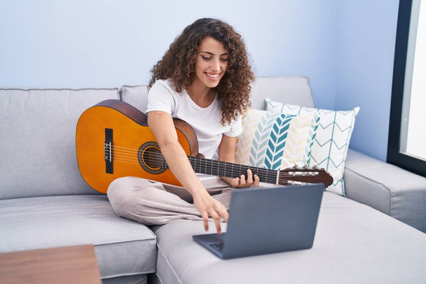 Young beautiful hispanic woman having online classical guitar class sitting on sofa at home - Фото, изображение
