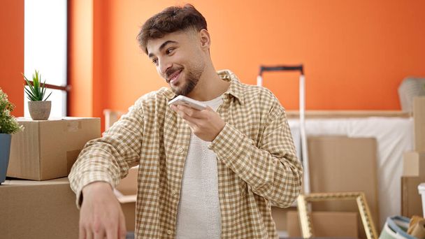 Young arab man smiling confident sending voice message by smartphone at new home - 写真・画像