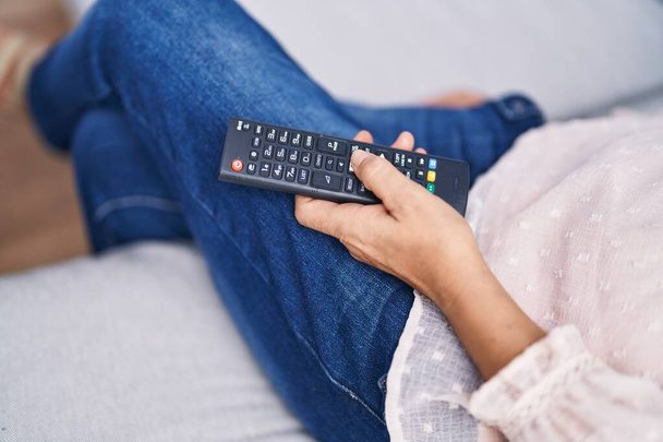 Жінка середнього віку з сірим волоссям тримає телевізор пульт дистанційного керування, сидячи на дивані вдома
 - Фото, зображення