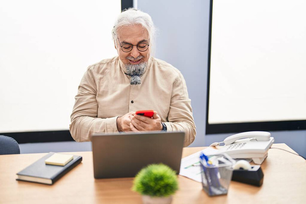 Homme d'affaires aux cheveux gris d'âge moyen utilisant un ordinateur portable et un smartphone au bureau - Photo, image