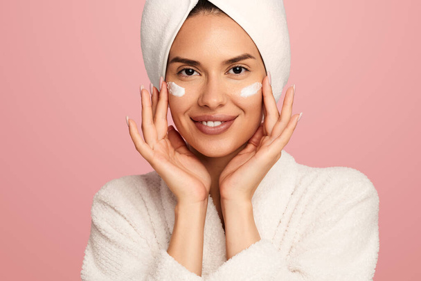 Smiling young female model in soft bathrobe and towel on head applying moisturizing cream on cheeks while standing against pink background - Foto, afbeelding