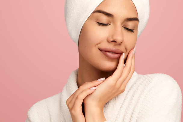 Young tender female in white bathrobe with towel on head and closed eyes touching face gently while standing against pink background after spa procedure - Foto, afbeelding