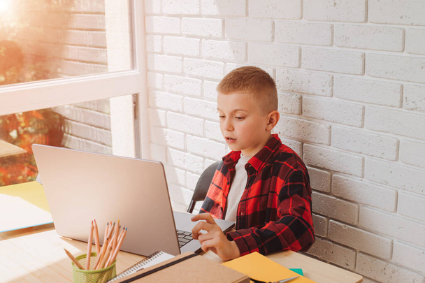 Een lachende schoolgaande jongen zit aan een tafel met een laptop. Kinderen en gadgets. Vooraanzicht - Foto, afbeelding