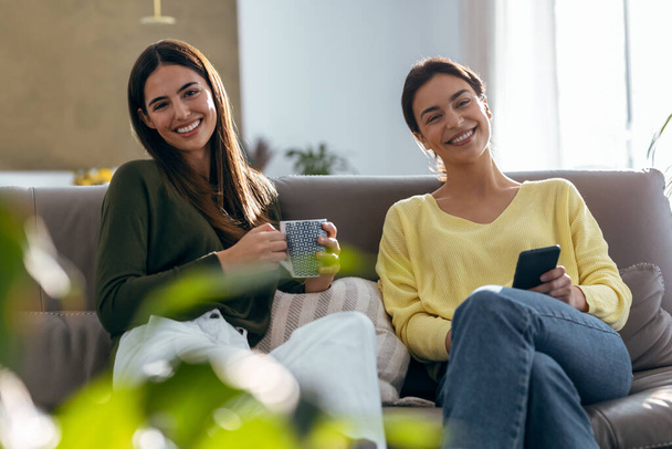 Shot of two smiling young women talking while drinking coffee sitting on couch in the living room at home. - Фото, зображення