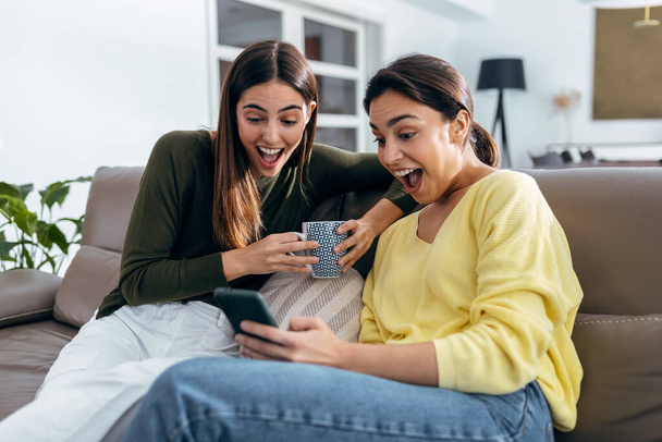 Shot of two smiling young women talking while watching smartphone sitting on couch in the living room at home. - Foto, Imagem