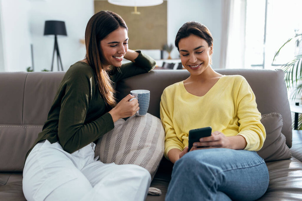 Shot of two smiling young women talking while watching smartphone sitting on couch in the living room at home. - Fotó, kép