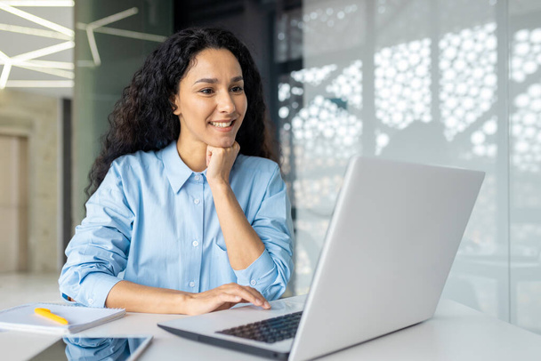Happy and smiling hispanic businesswoman typing on laptop, office worker with curly hair ,happy with achievement results, at work inside office building - Photo, Image
