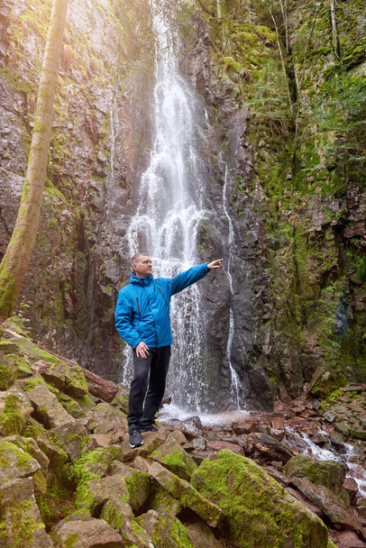 Tourist attraction of Germany - falls of Burgbach Waterfall near Schapbach, Black Forest, Baden-Wurttemberg, Germany. Man hiker in blue jacket standing on stone and looks at flow of falling water - Fotó, kép