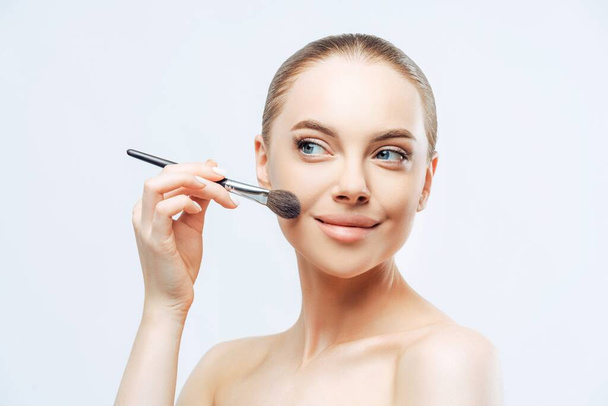 Facial treatment, cosmetology. Young woman, dark hair, applies powder with brush, sideways, naked body - Photo, Image