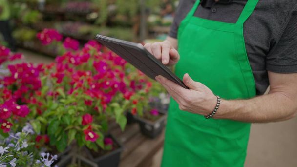 Close-up of staff_s hand holding tablet browsing inventory inside local business. Young man wearing green apron using modern technology at Flower Shop - Fotografie, Obrázek