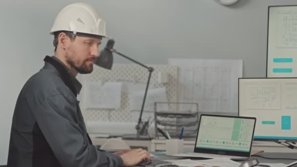 Lento medio de sonriente ingeniero de diseño masculino caucásico en sombrero blanco mirando a la cámara mientras está sentado en el escritorio de la oficina con esquemas de tecnología en monitores de computadora - Metraje, vídeo