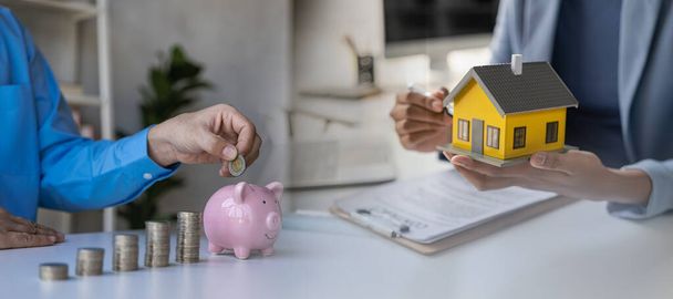 Coins in piggy bank with wooden house on table to save money for house purchase, financial concept for savings and borrowing. Horizontal cross section. - 写真・画像