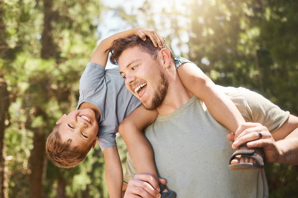 Son, father and back in outdoors with portrait for happiness for bonding and fun in summer on the weekend. Adventure, man and boy on shoulders with smile in nature for childhood with love and care - Photo, Image