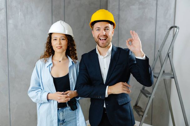 We've got the go ahead to renovate. Attractive young contractor and customer standing in room in helmet and looking at camera. - Zdjęcie, obraz