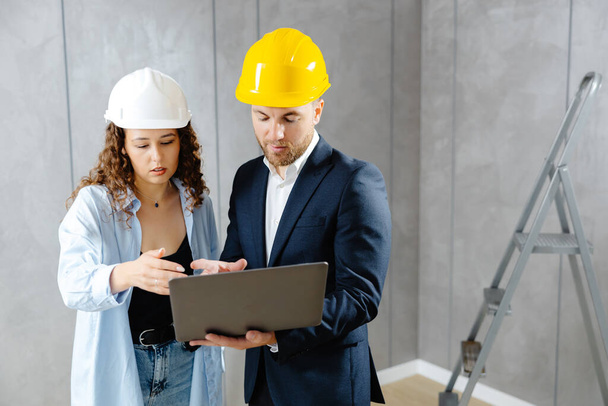 Attractive young woman contractor is talking to customer in helmet, and demonstrates sequence of repairs in living room on laptop. Meeting of landman of apartment with engineer or architect - Fotó, kép