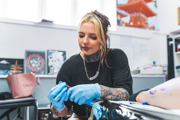 ένα νεαρό κορίτσι καλλιτέχνης τατουάζ προετοιμασία ένα μηχάνημα τατουάζ για να αρχίσει σχέδιο, μέτρια βολή, σαλόνι τατουάζ. Υψηλής ποιότητας φωτογραφία - Φωτογραφία, εικόνα