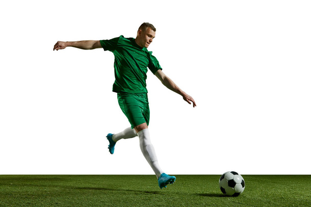 Young man in green uniform, football in motion, playing, kicking ball on sports field against white background. Concept of professional sport, action, lifestyle, competition, hobby, training, ad - Фото, изображение