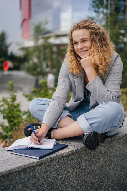 Nuori naisyrittäjä älykkäässä satunnaisasussa istuu rajalla ja kirjoittaa muistikirjaan työskennellessään projektissa kaupungissa - Valokuva, kuva