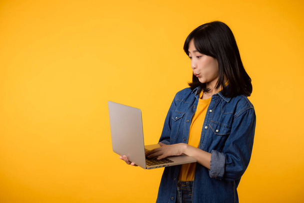 portret gelukkig jonge vrouw dragen geel t-shirt en denim shirt met laptop en gebruik notebook geïsoleerd op gele studio achtergrond. bedrijfstechnologie communicatieconcept. - Foto, afbeelding