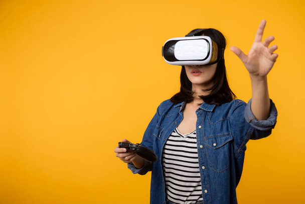 Ázsiai fiatal nő boldog siker visel virtuális valóság headset játék szórakoztató és gazdaság joystick vezérlő elszigetelt sárga háttér. A kiber jövő videojáték koncepciójának nyertese. - Fotó, kép