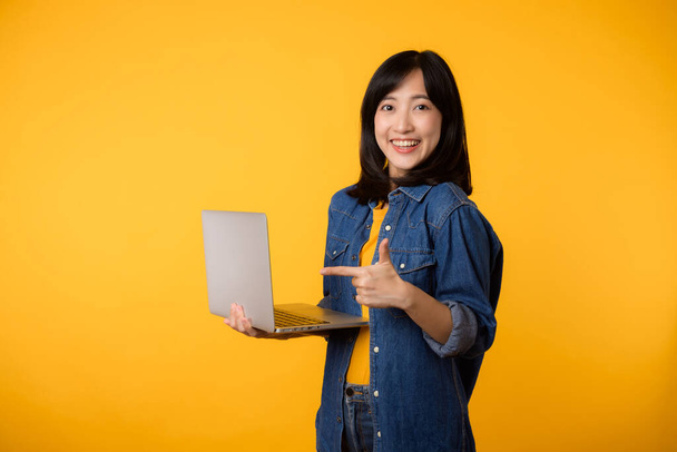 portrait happy young woman wearing yellow t-shirt and denim shirt holding laptop and point finger to screen isolated on yellow studio background. business technology application communication concept. - Photo, Image