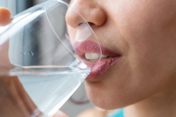 Close up of beautiful sporty woman drinking a glass of water while standing near the window at home - Foto, afbeelding