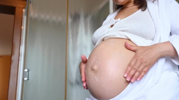 Midsection of a gravid expectant woman massaging her pregnant belly, frotando estrías cosméticos en el baño en casa. Procedimientos caseros de spa, cuidado de la piel y el cuerpo en tiempo de embarazo. Concepto de higiene - Metraje, vídeo