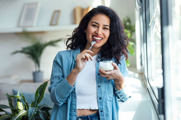Shot of happy beautiful woman eating yogurt while standing in living room at home. - Фото, зображення