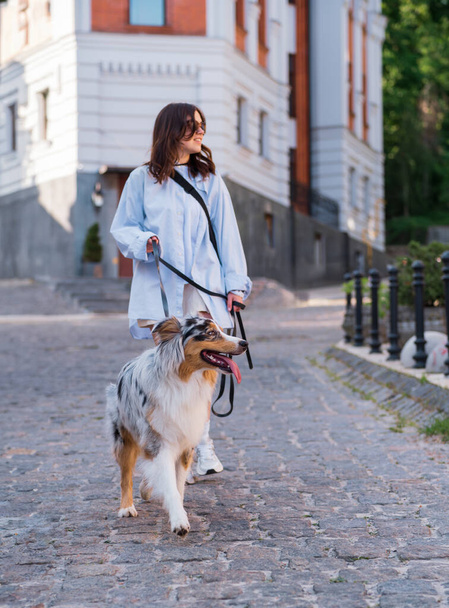 Mujer joven y su perro pastor asutraliano en la correa, enfoque selectivo en el perro. Caminar con mascotas en el centro de la ciudad, imagen de estilo de vida - Foto, Imagen