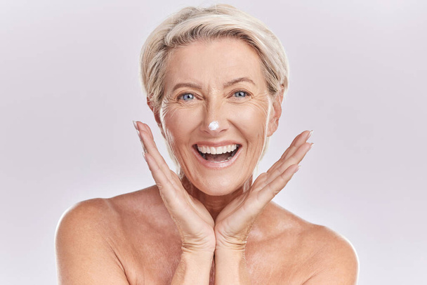 Skincare, portrait and mature woman with cream on face for anti aging, dermatology and collagen on white background. Facial, happy senior model with lotion or sunscreen in studio with beauty product - Foto, Imagen