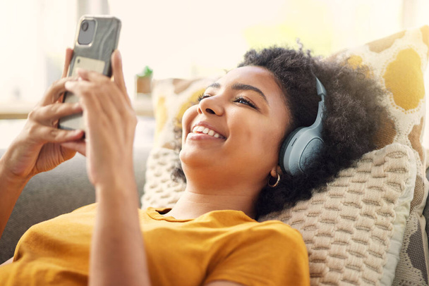 Relax, couch and woman with a smartphone, headphones and home with happiness, stress relief and streaming music. Smile, female person or girl on a sofa, headset and chilling with a cellphone and song. - Fotó, kép