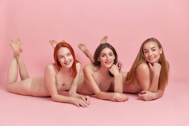 Relaxation and well-being. Beautiful redhead, brunette and blonde young women lying on floor in underwear against pink studio background. Concept of beauty, body and skin care, health, spa, ad - Zdjęcie, obraz