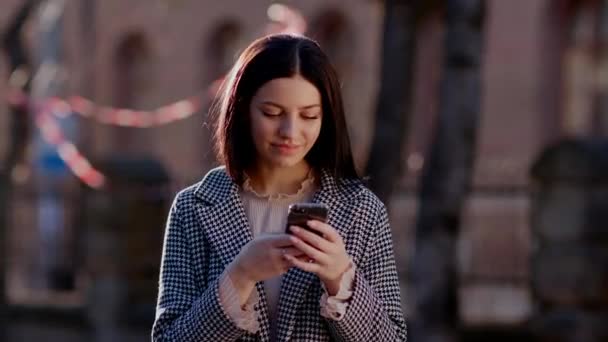 Een leuk meisje staat op straat en typt aan de telefoon. Het meisje staat tegen de achtergrond van gebouwen en bomen. Hoge kwaliteit 4k beeldmateriaal - Video