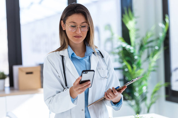 Aufnahme einer schönen Ärztin, die medizinische Dokumente überprüft, während sie während der ärztlichen Beratung das Handy im Stehen benutzt. - Foto, Bild