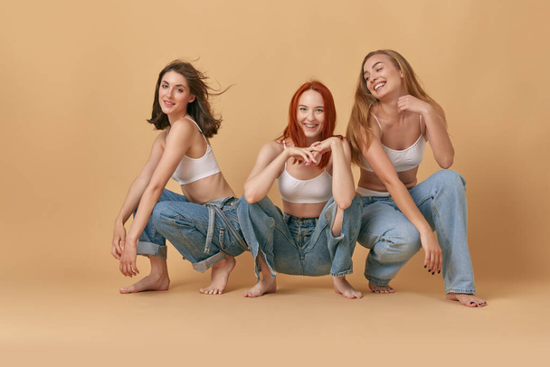 Portrait of three beautiful young brunette, redhead and blonde women in white top and jeans posing against studio background. Concept of beauty, body and skin care, health, fitness, wellness, ad - Photo, image
