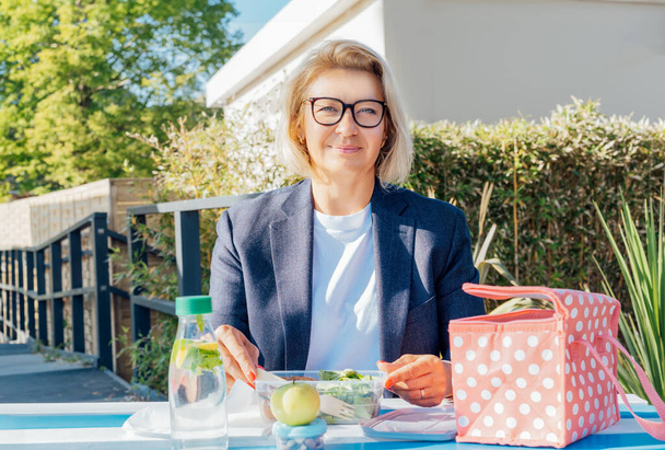 Portrait of smiling middle-aged business woman having lunch at the park outdoor cafe, picnic area during her break. Take away balanced diet lunch box with salad. Healthy eating habits and well-being. - Photo, Image