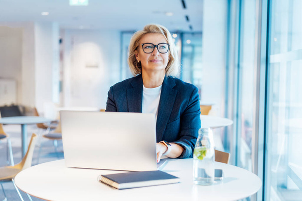 50s confident mature businesswoman dreamy looking at window, middle-aged experienced senior female professional working on laptop in open space office. Female entrepreneur working remotely. - Foto, imagen