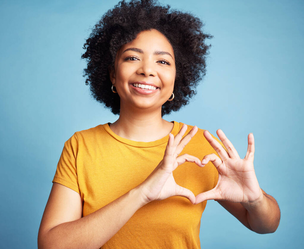 Happy, portrait and woman with a heart shape for romance, care or flirting expression in a studio. Happiness, smile and young female model with love gesture, emoji or sign isolated by blue background. - Zdjęcie, obraz