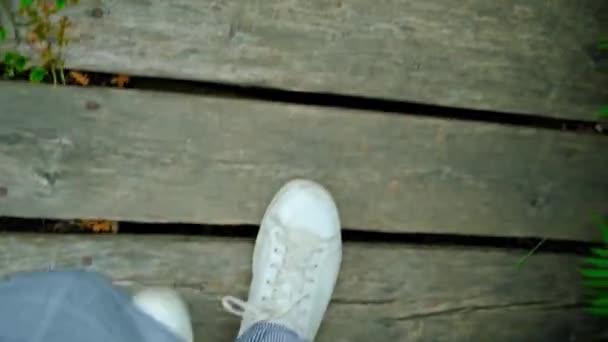 Donna in sneakers bianche che cammina su assi di legno lungo uno stretto sentiero nel giardino botanico, gambe in primo piano vista dall'alto - Filmati, video