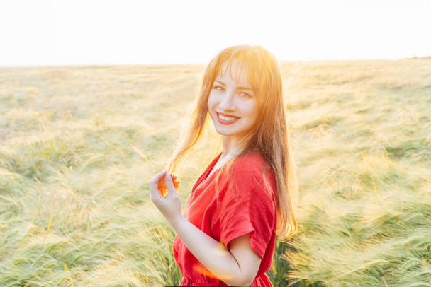 Mladá šťastná žena v červených šatech kráčí v pšeničném poli při západu slunce. Dýchej svobodně. Pozitivní emoce pociťující život, klid mysli. Duševní zdraví. Relaxace přírody. Měkké selektivní zaměření. - Fotografie, Obrázek