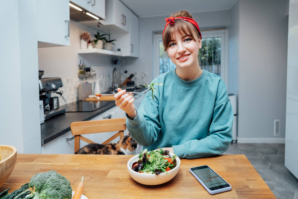Smiling young with woman with opened daily veganuary calendar on phone app while eating fresh green salad on the wooden table. Healthy vegetarian and vegan food diet planning. Selective focus. - 写真・画像