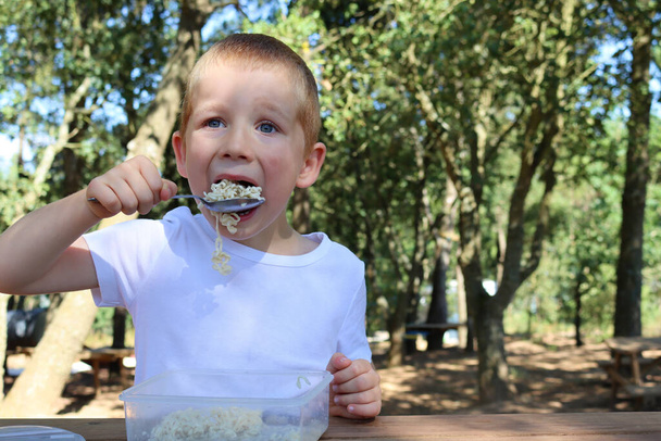 A hungry blond boy brings a spoonful of noodles to his mouth. Six year old boy having lunch in the park on a summer day - Photo, Image