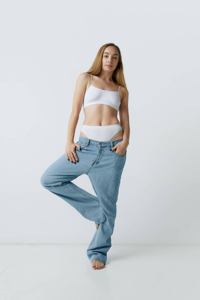 Full-length portrait of beautiful young blonde woman with slim body posing in jeans and top against grey studio background. Concept of beauty, body and skin care, health, fitness, wellness, ad - Zdjęcie, obraz