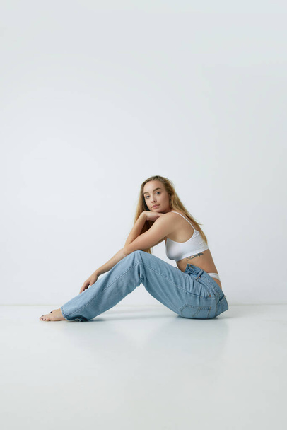 Tenderness. Beautiful young blonde woman with slim body posing in white top and jeans against grey studio background. Concept of beauty, body and skin care, health, fitness, wellness, ad - Valokuva, kuva