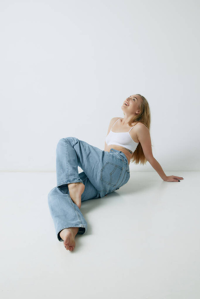 Beautiful, smiling, young blonde woman with slim body posing in white top and jeans against grey studio background. Concept of beauty, body and skin care, health, fitness, wellness, ad - Φωτογραφία, εικόνα