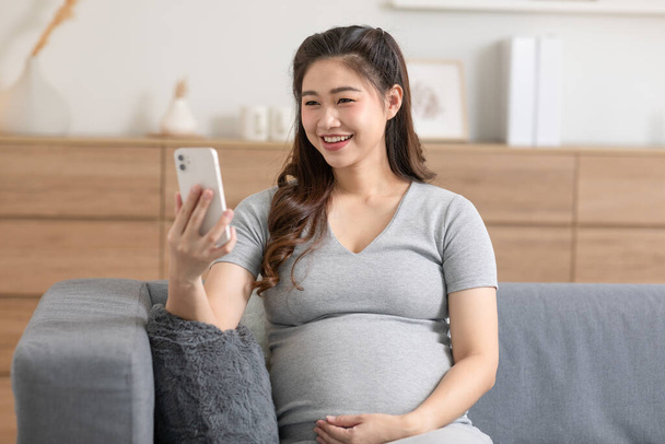 Happy pregnant asian woman use mobile phone to watch video media and relax with social media online.Pregnant woman smile and laughing lying on couch resting at home.Pregnancy and Technology Concept - 写真・画像